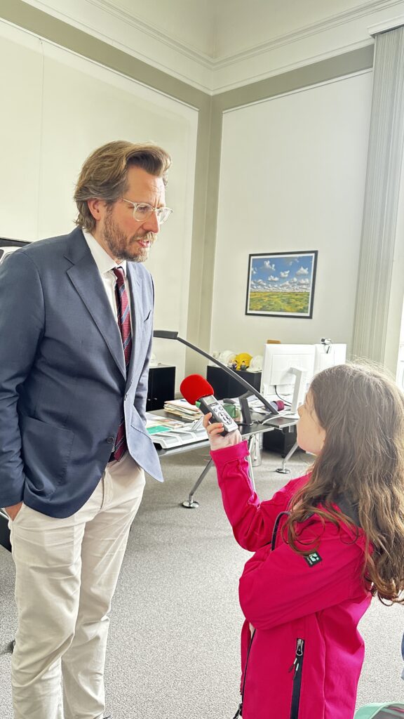 Links steht Christian Budde mit weißer Leinenhose, blauem Jacket und Krawatte und spricht in ein Aufnahmegerät, dass ihm von einem Mädchen mit langen braunen Haaren und einer pinken Softshell-Jacke entgegen gestreckt wird. Im Hintergrund steht ein voller Schreibtisch.