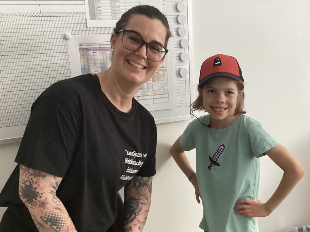 Foto eines Kindes mit rot-schwarzer Kappe und grünem T-Shirt neben einer Frau mit dunklen Haaren einer Brille und einem dunklen T-Shirt.