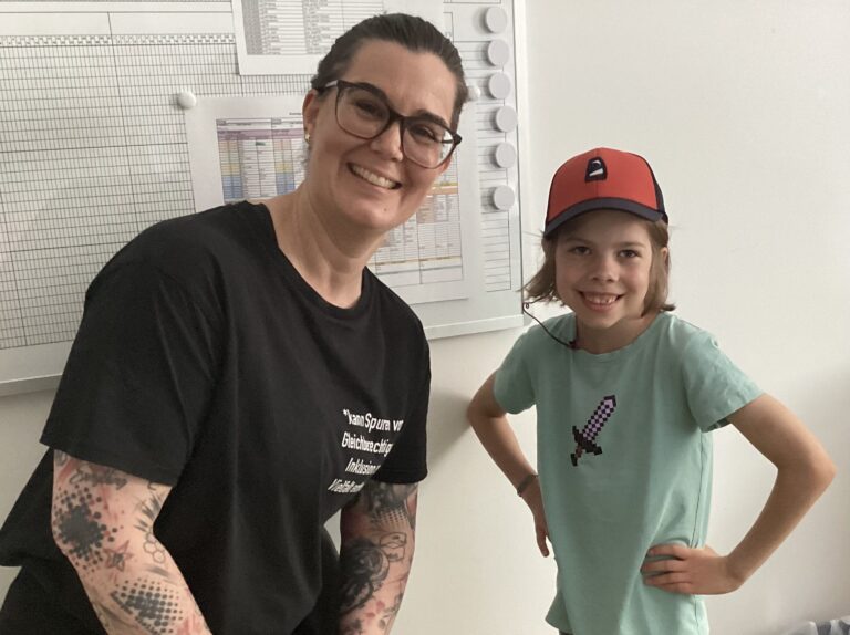 Foto eines Kindes mit rot-schwarzer Kappe und grünem T-Shirt neben einer Frau mit dunklen Haaren einer Brille und einem dunklen T-Shirt.