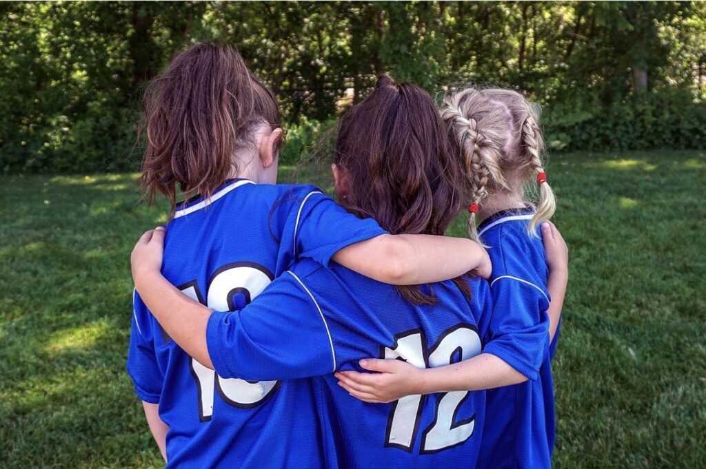 Drei Mädchen mit blauen Fußballtrikots mit weißen Nummern von hinten. das rechte Mädchen hat zwei blonde geflochtene Zöpfe mit roten Haargummis, das mittlere Mädchen trägt einen braunen Pferdeschwanz und das linke Mädchen auch. Sie stehen draußen auf einer Rasenfläche und im Hintergrund sind Bäume und Büsche.