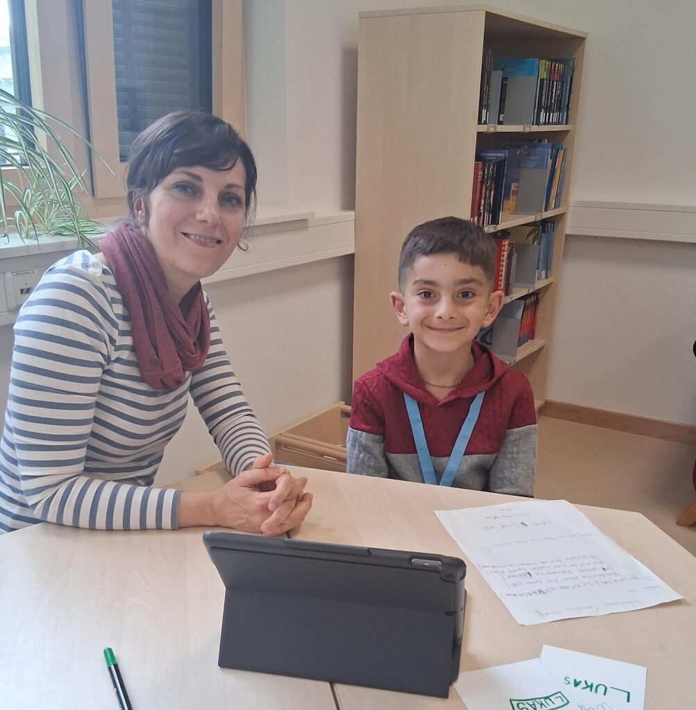 Christina Wolff und Kinder-Reporter Lukas sitzen an einem Tisch und lächeln in die Kamera. Vor ihnen auf dem Tisch liegen Notizen, ein Stift und ein iPad.